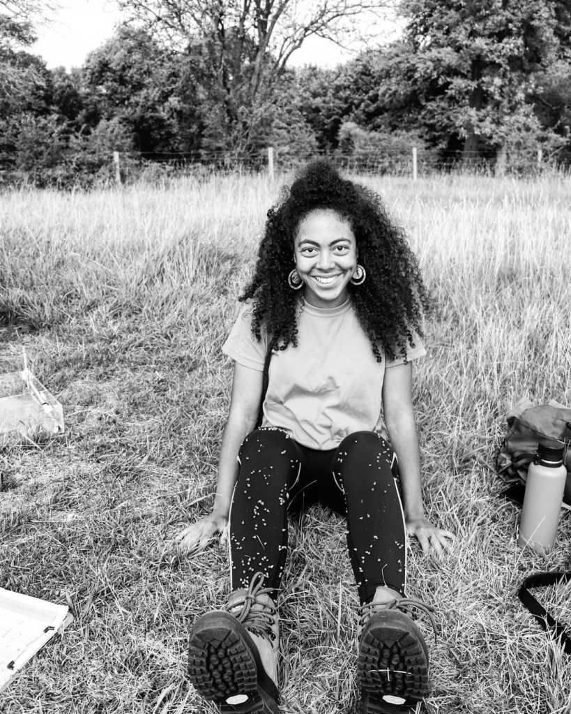  A black and white photo of Sara Middleton, she has long curly hair and is wearing round earings, leggings and a t-shirt and is sitting in the grass smiling and covered in seed pods.