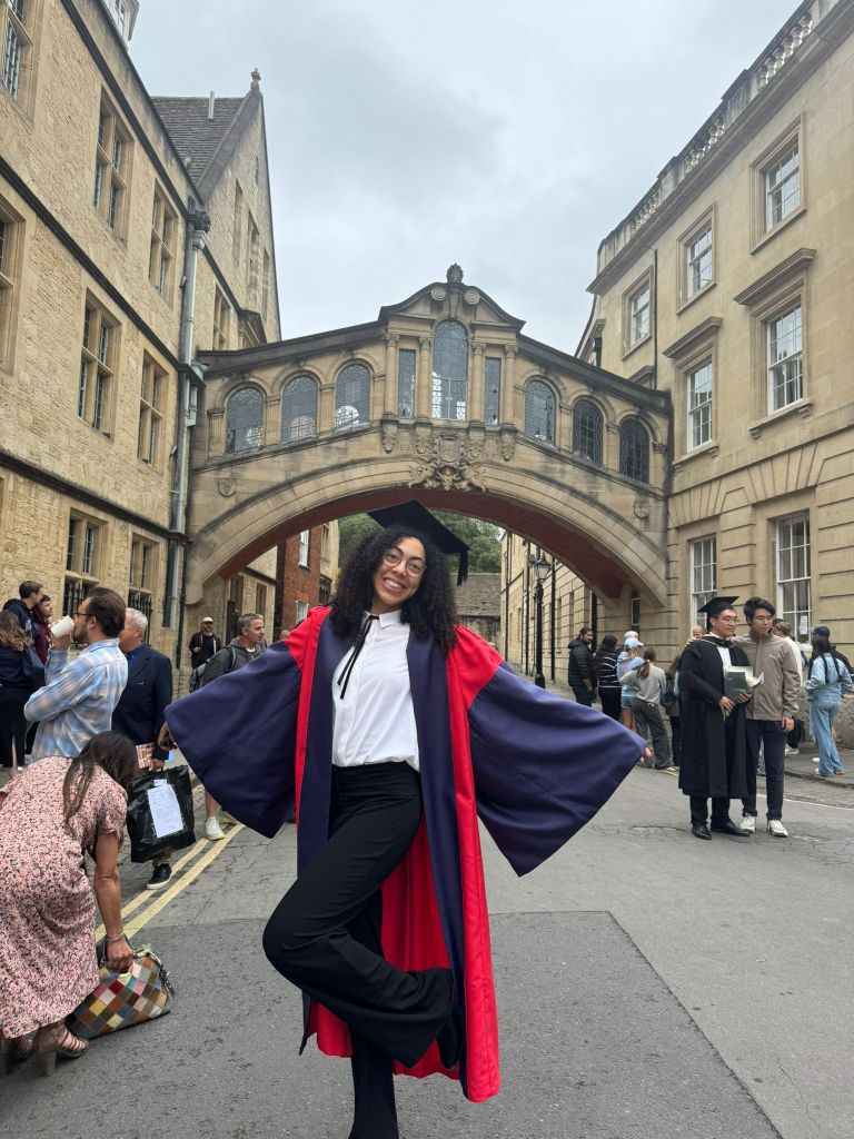 photo of Sara in academic dress celebrating her DPhil graduation in front of Hertford bridge